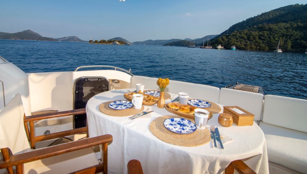 A beautifully set dining table on the shaded aft deck of a luxury motor yacht, overlooking a pristine mediterranean bay.