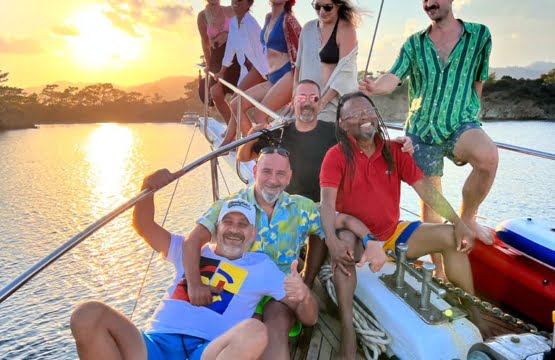 A wide-beam traditional wooden gulet anchored in the crystal-clear waters of a pine-fringed gocek bay, with a family relaxing on the spacious aft deck.