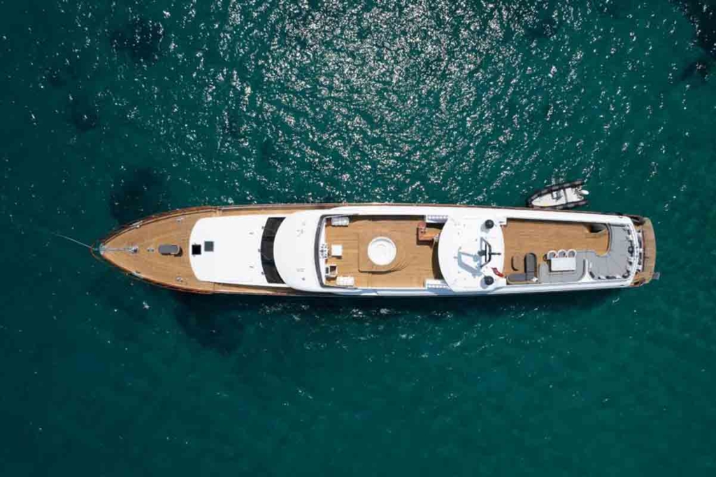 Luxury Motor Yacht Fly Bridge With Jaccuizi | Yacht Charter &Amp; Bareboat Rental Direct From Owners | Yachttogo Aerial view of a luxury motor yacht flybridge featuring a deck jacuzzi, spacious lounging areas, and a tender boat, cruising through clear turquoise waters.