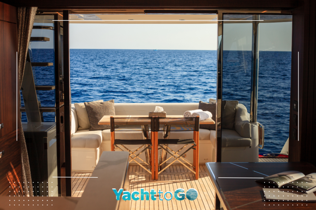 Interior view from a luxury motor yacht galley looking out through open glass doors to a teak wood aft deck with a dining table, chairs, and a wide-angle view of the deep blue ocean horizon.